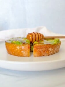 A slice of toasted bread topped with creamy avocado and drizzled honey sits on a white plate—a simple presentation inspired by the Best Avocado Toast Recipe, with a honey dipper resting on top.
