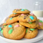 A plate of chocolate chip cookies topped with pastel-colored candy-coated chocolates, made using our Apple Cookies Recipe, sits beside a glass of milk in the background.