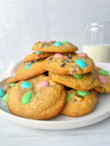 A plate of chocolate chip cookies topped with pastel-colored candy-coated chocolates, made using our Apple Cookies Recipe, sits beside a glass of milk in the background.