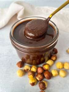 A glass jar filled with homemade Nutella chocolate hazelnut spread, with a spoon dipped in it. Whole hazelnuts are scattered around the jar on a light-colored surface.
