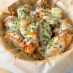 A basket lined with parchment paper filled with crispy Garlic Parmesan Wings, each covered in a creamy, white garlic parmesan sauce and garnished with chopped herbs.