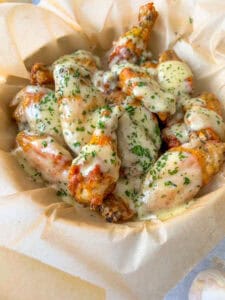 A basket lined with parchment paper filled with crispy Garlic Parmesan Wings, each covered in a creamy, white garlic parmesan sauce and garnished with chopped herbs.