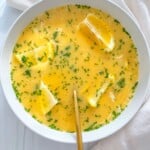 A bowl of yellow soup inspired by modern nonna recipes is garnished with parsley and lemon wedges, with a gold spoon resting inside. The bowl sits on a white surface beside a cream-colored cloth.