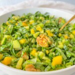 A bowl of fresh green salad, similar to a vibrant Mango Salad, featuring chopped spinach, cucumbers, yellow bell peppers, and walnuts, with a gold-colored fork resting inside.