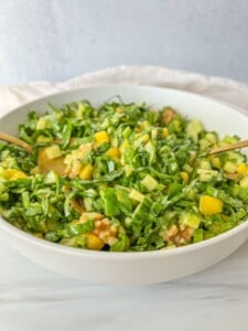 A white bowl filled with a fresh Peach Burrata Salad containing chopped greens, diced yellow bell peppers, cucumber, and walnuts, with two serving utensils inside.