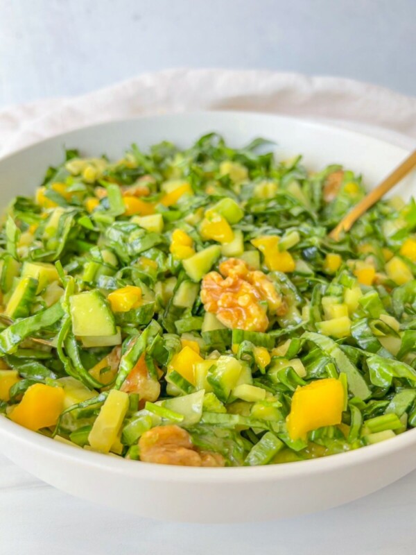 A bowl of fresh green salad, similar to a vibrant Mango Salad, featuring chopped spinach, cucumbers, yellow bell peppers, and walnuts, with a gold-colored fork resting inside.
