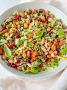 A white bowl filled with a colorful bean salad, including kidney beans, cannellini beans, and pinto beans, mixed with chopped cucumbers, red onions, fresh herbs, and basil leaves.
