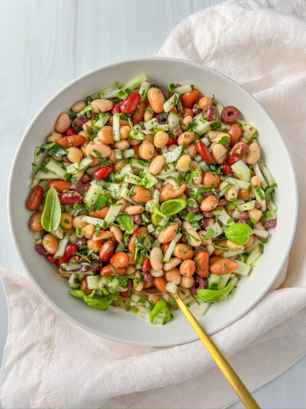 A white bowl filled with a vibrant Mixed Bean Salad of chopped cucumber, red onion, corn, fresh herbs, and basil leaves rests on a light cloth with a golden fork inside the bowl.