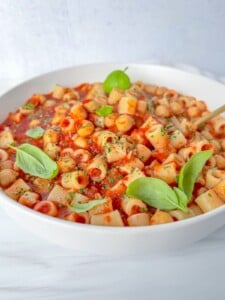 A white bowl filled with ditalini pasta in tomato sauce, garnished with chopped herbs and fresh basil leaves, sits on a white surface with a gold spoon—perfect alongside a crispy air fryer chickpeas for a high-protein snack.