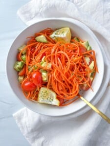A white bowl filled with spiralized carrots, cherry tomatoes, avocado chunks, and lime wedges—reminiscent of The Kourtney Kardashian Salad—set on a white napkin with a gold fork.