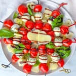 A plate of Valentines Appetizer caprese skewers with cherry tomatoes, mozzarella balls, basil leaves, and heart-shaped cheese slices, drizzled with balsamic glaze, arranged beautifully on a white dish.