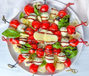 A plate of Valentines Appetizer caprese skewers with cherry tomatoes, mozzarella balls, basil leaves, and heart-shaped cheese slices, drizzled with balsamic glaze, arranged beautifully on a white dish.
