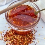 A glass jar filled with chili-infused honey, topped with a whole dried chili and a honey dipper resting on the rim. Crushed red pepper flakes are scattered around the jar on a white surface.