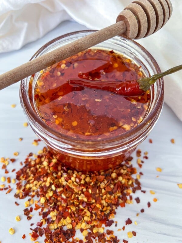 A glass jar filled with chili-infused honey, topped with a whole dried chili and a honey dipper resting on the rim. Crushed red pepper flakes are scattered around the jar on a white surface.