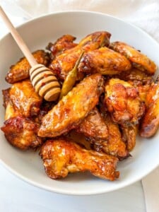 A bowl of glazed chicken wings topped with a whole chili pepper and drizzled honey, with a honey dipper resting on the side of the dish.