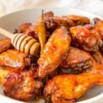 A white bowl filled with glazed chicken wings, topped with honey drizzled from a honey dipper, and garnished with a whole chili pepper.