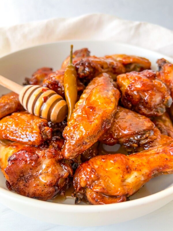 A white bowl filled with glazed chicken wings, topped with honey drizzled from a honey dipper, and garnished with a whole chili pepper.