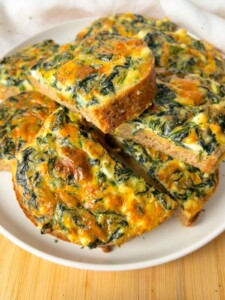Slices of baked spinach and cheese frittata are arranged on a white plate, showing a golden-brown crust and a colorful mixture of spinach and melted cheese inside.