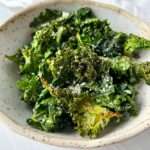 A bowl of crispy Air Fryer Kale Chips sprinkled with grated cheese, served in a speckled ceramic dish on a white surface.