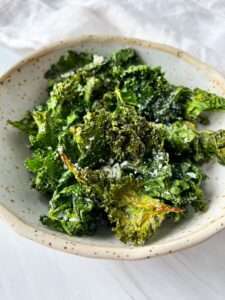 A bowl of crispy Air Fryer Kale Chips sprinkled with grated cheese, served in a speckled ceramic dish on a white surface.