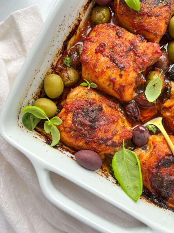 A white baking dish filled with Pizza Sauce Baked Chicken thighs, surrounded by green and black olives, fresh basil leaves, and garnished with herbs. The dish rests on a white cloth.