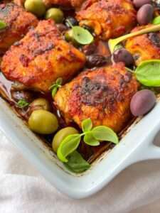 A white baking dish with golden roasted chicken thighs in pizza sauce, garnished with green and purple olives and fresh basil leaves.
