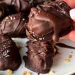 A hand holds a chocolate-covered date over a plate filled with similar chocolate-coated sweets, all drizzled with chocolate and sprinkled with small crumbs.