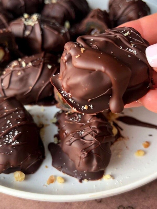 A hand holds a chocolate-covered date over a plate filled with similar chocolate-coated sweets, all drizzled with chocolate and sprinkled with small crumbs.