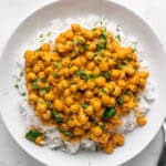 A white plate filled with a hearty serving of chickpea curry on top of fluffy white rice. The dish is garnished with fresh green herbs, adding a touch of color. A small bowl of herbs is visible in the top left corner.