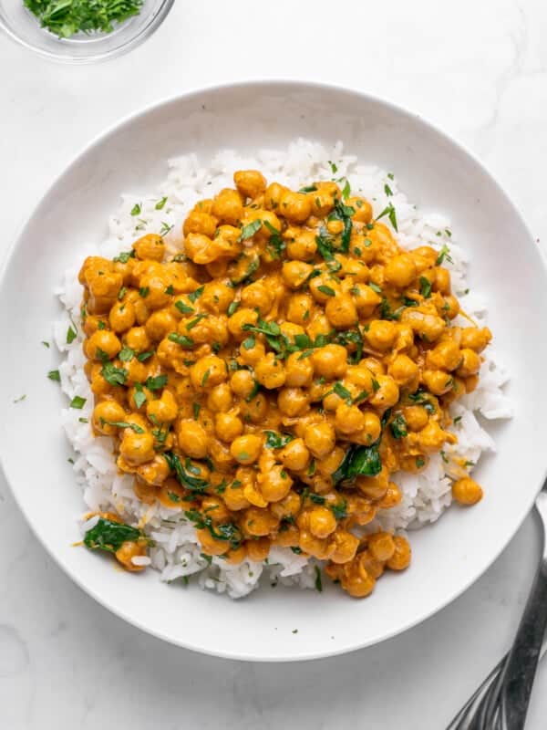 A white plate filled with a hearty serving of chickpea curry on top of fluffy white rice. The dish is garnished with fresh green herbs, adding a touch of color. A small bowl of herbs is visible in the top left corner.