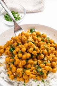 A plate of white rice topped with chickpea curry garnished with chopped cilantro. A fork is scooping the curry. In the background, a small bowl of fresh cilantro is visible on a white surface.