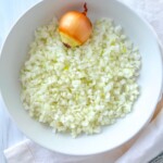 A white bowl filled with finely chopped onions showcases the results of learning How To Dice Onion, with a whole unpeeled onion placed on top and resting on a white cloth.