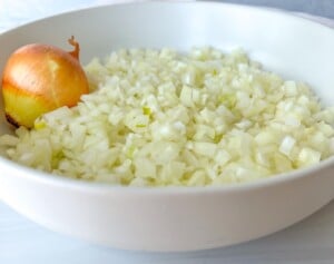 A white bowl filled with finely chopped onions and a whole unpeeled onion resting on the side—perfect for learning kitchen basics like how to shred lettuce.