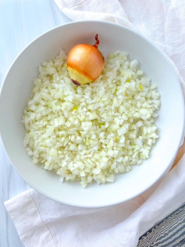 A white bowl filled with finely chopped onions showcases the results of learning How To Dice Onion, with a whole unpeeled onion placed on top and resting on a white cloth.