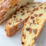 A loaf of golden brown Irish Soda Bread studded with raisins sits on a white surface, with two thick slices cut to reveal its soft, airy interior filled with more raisins.