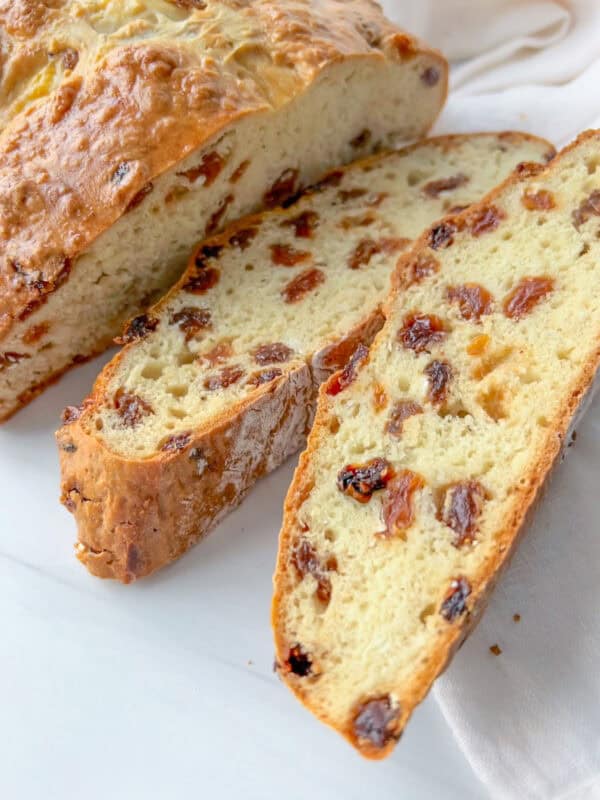 A loaf of golden brown Irish Soda Bread studded with raisins sits on a white surface, with two thick slices cut to reveal its soft, airy interior filled with more raisins.