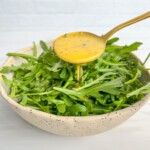 A spoon pours yellow salad dressing, inspired by a Hot Honey Recipe, over a bowl filled with fresh arugula leaves, set against a light background.