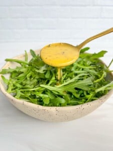 A spoon pours yellow salad dressing, inspired by a Hot Honey Recipe, over a bowl filled with fresh arugula leaves, set against a light background.