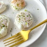 A white plate with three poached eggs sprinkled with herbs and black pepper sits next to a gold fork. The bright, minimal background highlights this perfect example of how to poach eggs beautifully.