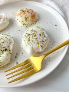 A white plate with three poached eggs sprinkled with herbs and black pepper sits next to a gold fork. The bright, minimal background highlights this perfect example of how to poach eggs beautifully.
