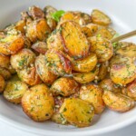 A white bowl filled with roasted baby potatoes, sliced and coated in fresh herbs and seasoning, topped with crumbled feta for a classic Greek Potatoes with Feta dish. A fork rests invitingly in the bowl.