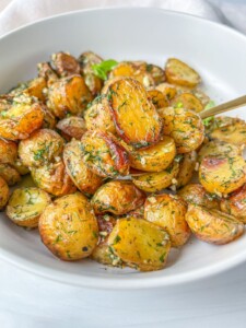A white bowl filled with roasted baby potatoes, sliced and coated in fresh herbs and seasoning, topped with crumbled feta for a classic Greek Potatoes with Feta dish. A fork rests invitingly in the bowl.