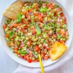 A bowl of chopped salad with tomatoes, cucumbers, onions, and herbs, garnished with fresh mint leaves. Two pieces of pita chips and two gold utensils are in the bowl, evoking the freshness of a Strawberry Spinach Salad on a white cloth.