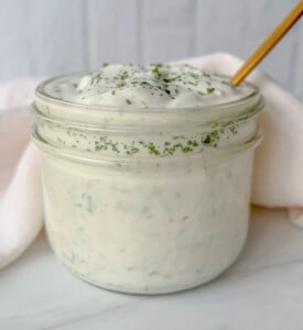 A glass jar filled with creamy white ranch dressing, topped with green herb flakes, sits on a white surface with a gold spoon and a soft white cloth in the background.