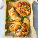 A tray lined with parchment paper holds mini pizzas topped with cheese and herbs, and several calzones. Fresh basil leaves are scattered on top for garnish.