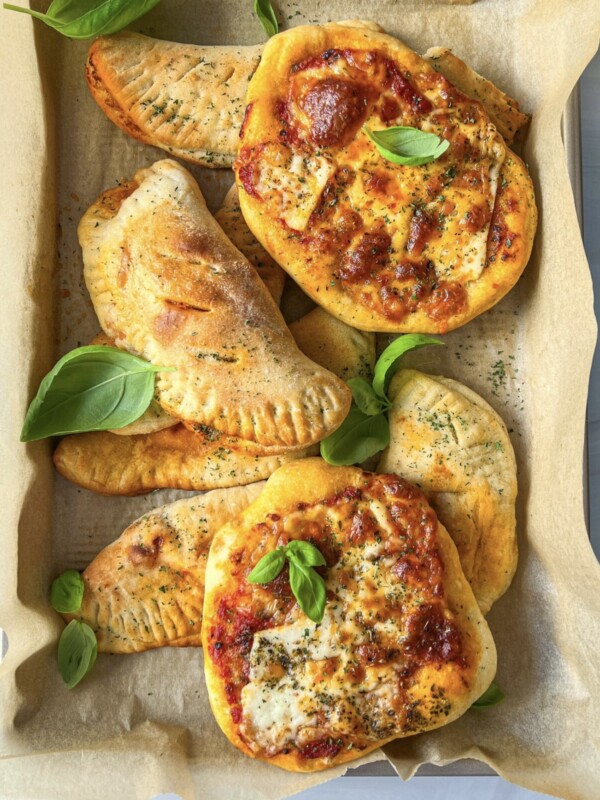 A tray lined with parchment paper holds mini pizzas topped with cheese and herbs, and several calzones. Fresh basil leaves are scattered on top for garnish.