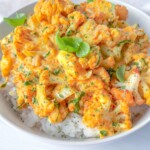 A bowl of white rice topped with roasted cauliflower coated in a creamy orange sauce, garnished with chopped herbs and fresh basil leaves.