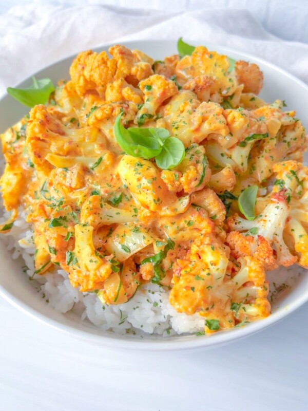 A bowl of white rice topped with roasted cauliflower coated in a creamy orange sauce, garnished with chopped herbs and fresh basil leaves.