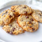 A white plate with several homemade Honey Almond Cookies made of oats, shredded coconut, and chocolate chips, set on a light-colored surface with a white cloth in the background.