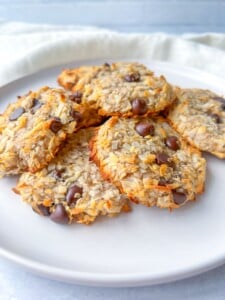 A white plate with several homemade Honey Almond Cookies made of oats, shredded coconut, and chocolate chips, set on a light-colored surface with a white cloth in the background.